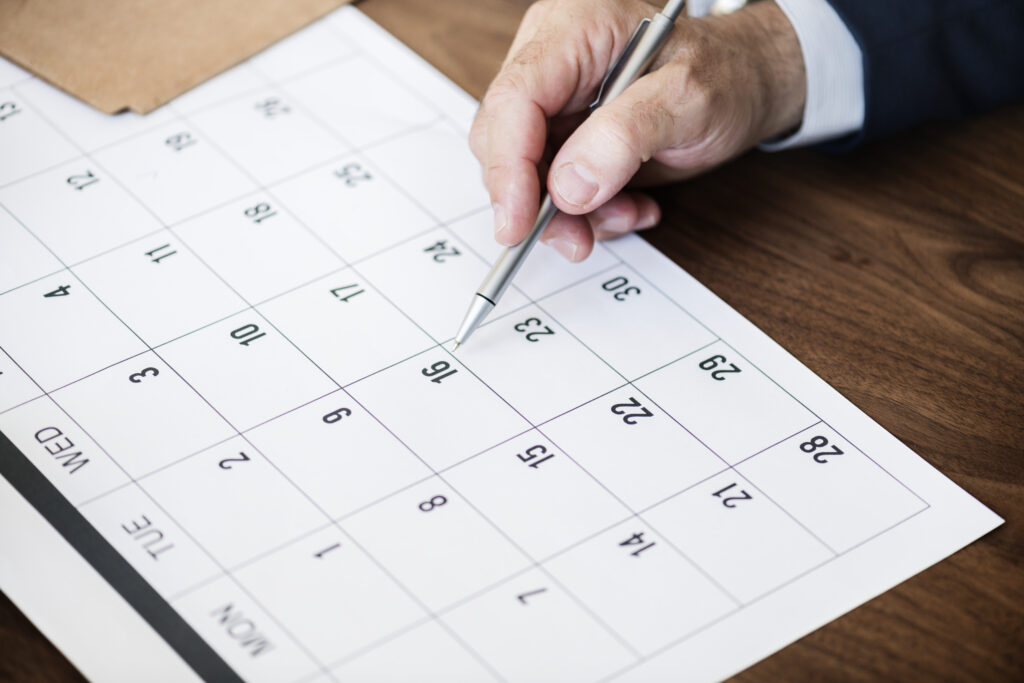 papel com calendário à mesa, mão do homem branco com caneta sobre a folha.